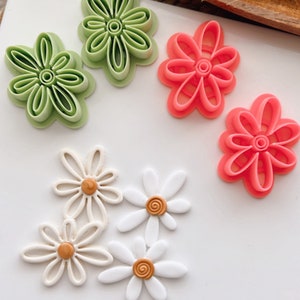May include: A set of five flower-shaped cookie cutters. Two are green, two are pink, and one is white with a brown centre.
