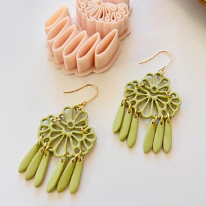 May include: A pair of light green, floral-shaped earrings with gold-colored hooks and dangling teardrop accents. A pink, flower-shaped clay cutter is in the background, suggesting a craft or jewelry-making theme.