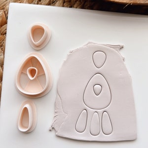 Puede incluir: Un juego de cortadores de galletas de plástico rosa en forma de mujer, corazón y tres formas de lágrima. Los cortadores están sobre una superficie blanca.