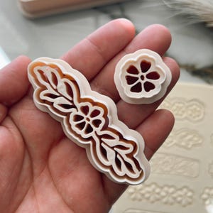 May include: A set of white plastic cookie cutters featuring floral and leaf designs. The cutters are held in a hand and are shown next to a sheet of white silicone with other cutters.