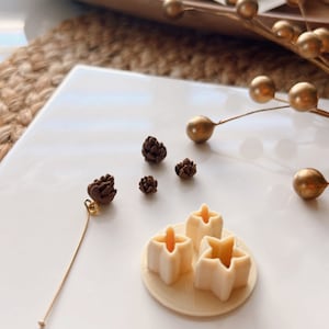 Puede incluir: Un plato de cerámica blanco con tres abalorios marrones en forma de piña y tres abalorios blancos en forma de estrella. Los abalorios están dispuestos en círculo en el plato. Un alambre dorado está unido a uno de los abalorios en forma de piña.