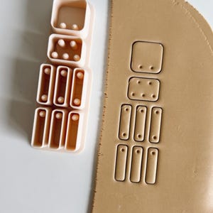 以下が含まれることがあります： 正方形、長方形、円形など、さまざまな形をしたピンクのプラスチック製のクッキーカッター。クッキー生地に装飾的な形を作るために使用されます。