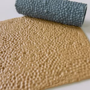 Puede incluir: Una lámina texturizada de color marrón claro con un patrón de pequeñas protuberancias en relieve. Un rodillo gris con una textura similar y llena de bultos descansa encima. El rodillo es cilíndrico y parece estar hecho de un material gomoso. Para manualidades o decoración.