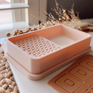 Puede incluir: Una bandeja de plástico rosa con una cuadrícula de pequeños agujeros y una sección más grande y lisa. La bandeja está sobre una superficie blanca junto a una estera de cuero marrón con tres recortes rectangulares.