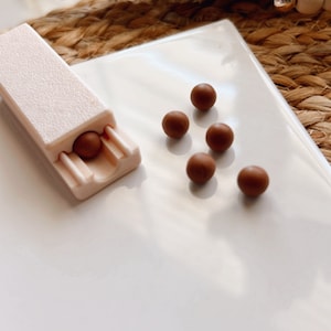 May include: A white plastic pill dispenser with a single brown pill inside. Six additional brown pills are scattered on a white surface.