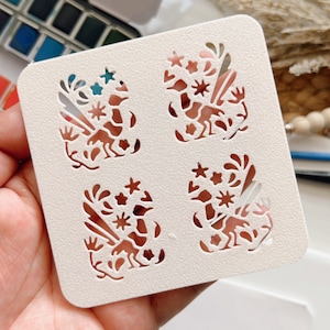 Puede incluir: Plantilla de plástico blanco con cuatro diseños recortados de pájaros estilizados con elementos florales. La plantilla se sostiene en una mano.