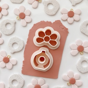 Op de afbeelding: Een set met benodigdheden voor het maken van oorbellen van polymer clay. De set bevat twee koekjesuitstekers, een in de vorm van een bloem en de andere in de vorm van een fles. Er zijn ook verschillende voorgevormde oorbellen van polymer clay in de vorm van bloemen en cirkels.