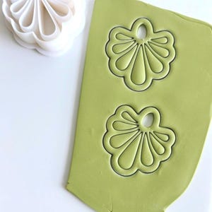 Puede incluir: Un cortador de pendientes de plástico blanco junto a un trozo de arcilla verde con dos formas de pendientes prensadas. La arcilla está sobre una superficie blanca. El cortador tiene forma de flor.
