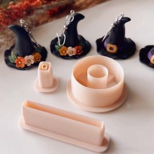May include: A set of four witch hat-shaped polymer clay earring molds, along with three pink plastic tools for shaping the clay. The molds are black, purple, and green with orange and white accents.
