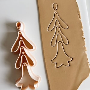 May include: A close-up of a cookie cutter in the shape of a flower with five petals. The cutter is made of white plastic and is sitting on a brown surface. The cutter is being used to cut out a flower shape from a piece of brown clay.