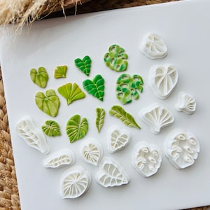 May include: A set of 18 leaf-shaped cookie cutters, including various shapes and sizes. The cutters are made of white plastic and are arranged on a white surface. Some of the cutters are green, representing the leaves.