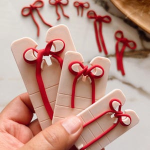 Puede incluir: Cuatro herramientas de plástico rosa con lazos de cinta roja para hacer lazos. Las herramientas son de diferentes tamaños y tienen ranuras para sujetar la cinta.