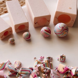 Può includere: Quattro stampi in silicone rosa per realizzare perline rotonde in pasta polimerica. Gli stampi sono di dimensioni diverse e sono mostrati con perline colorate a spirale realizzate in pasta polimerica.