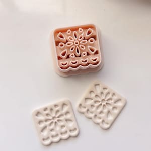 May include: Two white, decorative, square-shaped cookie cutters with a floral design. The cutters are on a white surface. A third cutter, which is closed, is in the background.