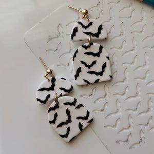Può includere: Orecchini bianchi e neri a tema pipistrello. Gli orecchini sono realizzati in un materiale bianco con un motivo a pipistrello nero. Gli orecchini hanno un perno e una sfera color oro. Sono disposti su una superficie bianca.