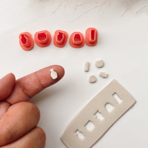 May include: A set of five pink plastic clay cutters shaped like different vases and bottles. The cutters are arranged in a row on a white surface. There are also several small pieces of white clay on the surface, and a larger piece of white clay with a rectangular shape and several vase-shaped cutouts.