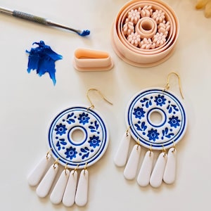 以下が含まれることがあります： 手作りの白と青の花柄イヤリング。ゴールドカラーのフックと白いしずく型のドロップが付いています。粘土ツールと粘土も写っています。イヤリングは製作中です。