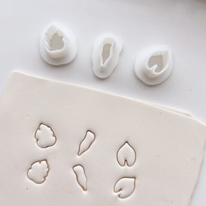 Puede incluir: Un juego de cortadores de arcilla polimérica blanca con varias formas de hojas, incluyendo una hoja grande, una hoja pequeña y una hoja de nenúfar. Los cortadores están dispuestos sobre una superficie blanca.