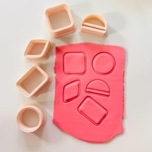 Puede incluir: Un conjunto de cortadores de galletas con formas geométricas de color rosa claro junto a un trozo de arcilla rosa con formas correspondientes presionadas. Incluye cuadrados, rectángulos, un círculo, un diamante y un semicírculo.