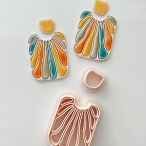 Puede incluir: Un conjunto de dos pendientes de cerámica con un diseño geométrico en tonos de amarillo, naranja, verde y azul. Los pendientes se muestran con sus correspondientes cortadores de arcilla.
