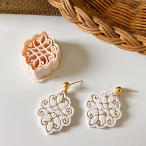 Puede incluir: Pendientes blancos y dorados con diseño floral y postes dorados, junto con un cortador de galletas a juego. Los pendientes y el cortador están sobre una superficie blanca, con una cesta de mimbre en el fondo.