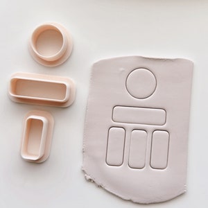 May include: A collection of light pink polymer clay cutters in various shapes, including a circle, rectangle, and rounded rectangles. A piece of clay with matching cut-out shapes is also present. The items are arranged on a white surface.