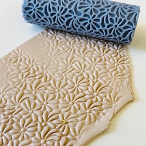May include: A gray floral pattern rolling pin next to a piece of clay with the same embossed floral design. The clay is a light tan color. The rolling pin is made of a hard material and is cylindrical in shape.
