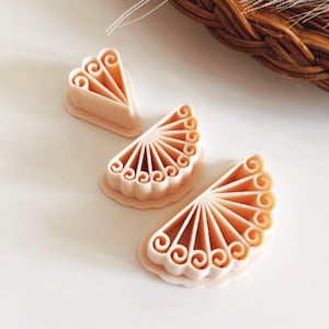 May include: Three light peach-coloured cookie cutters in a fan shape, arranged on a white surface. Each cutter has a scalloped edge and a detailed design of swirls and lines. The cutters are likely for baking or crafting.
