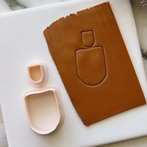 Puede incluir: Un juego de dos cortadores de galletas con forma de jarrón abstracto moderno. Los cortadores son de plástico blanco y se muestran sobre una superficie blanca con un trozo de arcilla marrón.