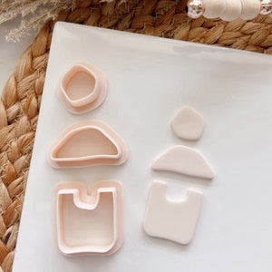 Puede incluir: Un juego de cuatro cortadores de galletas con forma de figuras geométricas abstractas. Los cortadores están hechos de plástico rosa claro y están dispuestos sobre una superficie blanca.