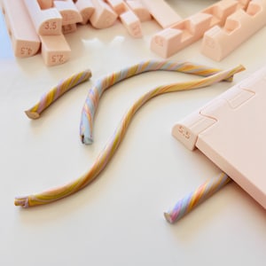 Puede incluir: Una colección de herramientas de arcilla polimérica rosa y bastones de arcilla coloridos. Las herramientas incluyen varias medidas en cm, y los bastones presentan un remolino de colores pastel. Los artículos están dispuestos sobre una superficie blanca.