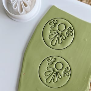 May include: A white plastic cookie cutter with a floral design sits beside a piece of green clay with two impressions of the same design. The clay is on a white surface.