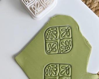 Pode incluir: Um cortador de argila polimérica branca e um pedaço de argila verde com um desenho floral em relevo. O cortador é quadrado com um padrão floral. A argila tem duas impressões do mesmo desenho. O fundo é uma superfície branca.