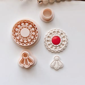 Puede incluir: Conjunto de cinco cortadores de galletas, que incluyen dos cortadores redondos con diseños intrincados, un cortador redondo con un centro rojo y dos cortadores con forma de flor.