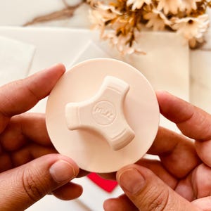 Puede incluir: Un tapón de botella rosa claro con un asa de tres puntas. El tapón tiene la palabra "m.r." impresa. El tapón se sostiene con dos manos. El fondo incluye una superficie blanca y un ramo de flores.