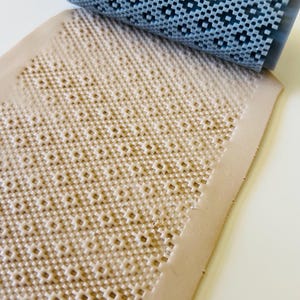 May include: A light gray textured roller rests on a beige, patterned surface. The roller has a grid-like design, and the surface has a repeating pattern of small, square indentations. The items appear to be craft tools.