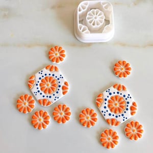 May include: A white plastic cookie cutter next to several orange flower-shaped decorations and two square decorations with orange and blue floral designs. The decorations are likely for baking or crafts.