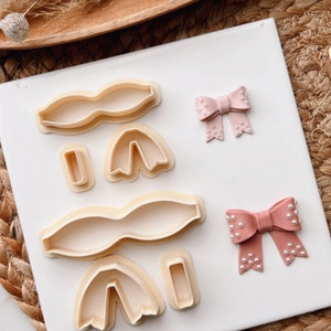 Puede incluir: Un juego de cinco cortadores de galletas con forma de lazos y tulipanes. Los cortadores están hechos de plástico blanco y están dispuestos sobre una superficie blanca. También se muestran dos lazos rosas con detalles de perlas.