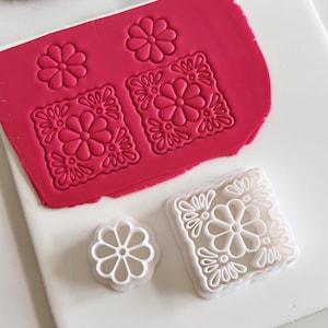 Puede incluir: Primer plano de herramientas y arcilla polimérica. Una lámina de arcilla rosa brillante con impresiones florales. También se ven dos cortadores de plástico blanco, uno redondo y otro cuadrado. Un salvamanteles de paja tejida con flecos coloridos está en el fondo.