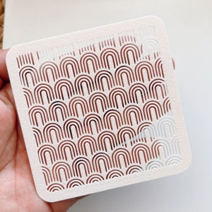 May include: White plastic stencil with a repeating pattern of brown arches. The arches are arranged in rows and create a geometric design.