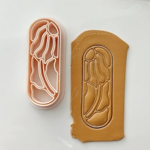 May include: A pink plastic cookie cutter with an abstract design is shown next to a piece of brown clay with the design impressed into it.