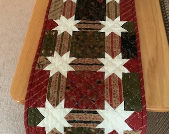 May include: A quilted table runner with a red, green, and white star pattern. The runner is made of fabric scraps in various patterns and colors.
