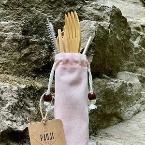Könnte beinhalten: Ein rosafarbenes wiederverwendbares Besteckset mit einer Holz-Gabel, einem Löffel, einem Messer und einem Strohhalm sowie einer Reinigungsbürste. Das Set befindet sich in einem rosafarbenen Kordelzugbeutel mit einem braunen Etikett, auf dem "Pauji Reduce Your Footprint" steht.