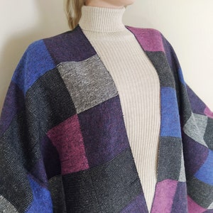 Puede incluir: Un chal con estampado de cuadros en tonos azul, morado, gris y negro. El chal está hecho de una tela suave y tejida y tiene un borde con flecos.