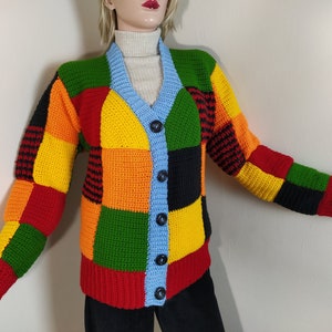 Könnte beinhalten: Ein bunter Häkelcardigan mit Patchwork-Muster in Rot, Orange, Gelb, Grün, Schwarz und Blau. Der Cardigan hat einen V-Ausschnitt und einen Knopfverschluss.