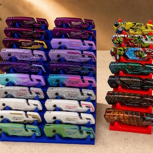 May include: A collection of multi-colored Milwaukee Fastback utility knives displayed in blue and red organizers. The knives feature various designs and colors, including purple, blue, white, and green. The image showcases a variety of utility knives.