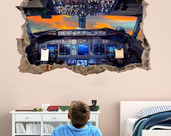 May include: A child sits on a green and white striped rug, looking up at a wall decal of an aircraft cockpit. The decal is designed to look like a hole in the wall, revealing the cockpit interior. The child is wearing a blue shirt and holding a football.