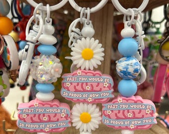 May include: Three keychains featuring white rings and pink brain-shaped charms with the text 'PAST YOU WOULD BE SO PROUD OF NOW YOU' in blue. The charms are accented with white daisy flowers, blue beads, and iridescent textured beads.