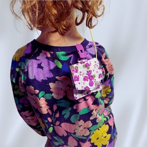 May include: A child wearing a black and purple floral print shirt with a small purple and white floral fabric pouch attached to the shirt with a purple clip.