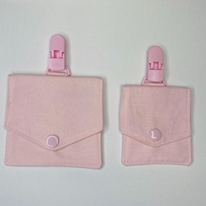 May include: Two pale pink fabric pacifier clips with a square pouch design. Each clip has a pink plastic clip at the top and a button closure on the front. The clips are in different sizes.